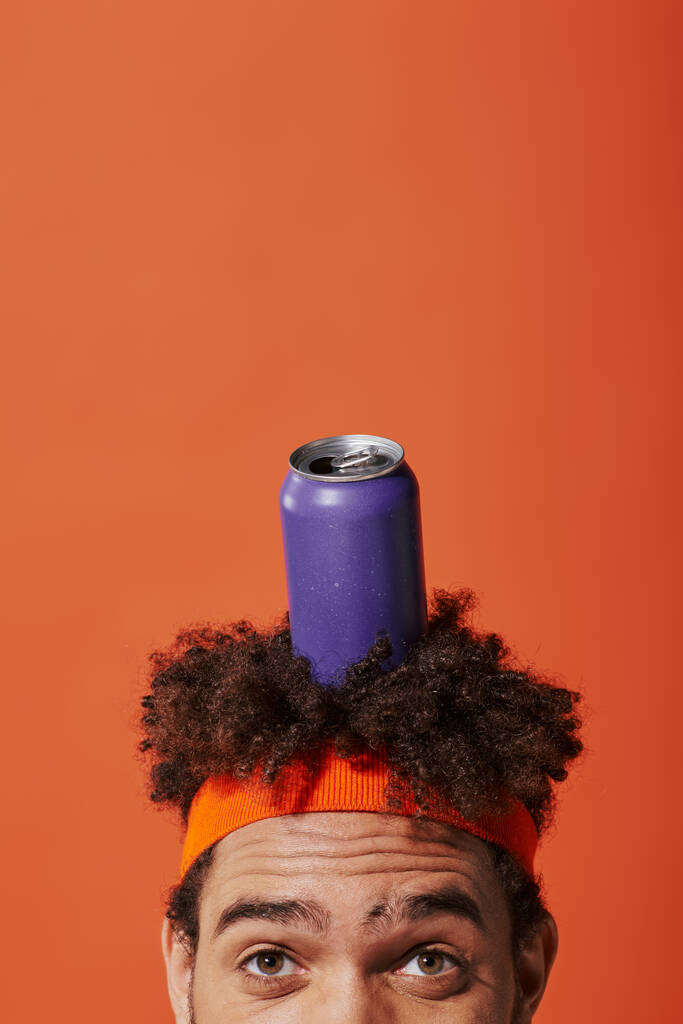 purple soda can on head of curly african american man with headband on orange background - Photo, Image