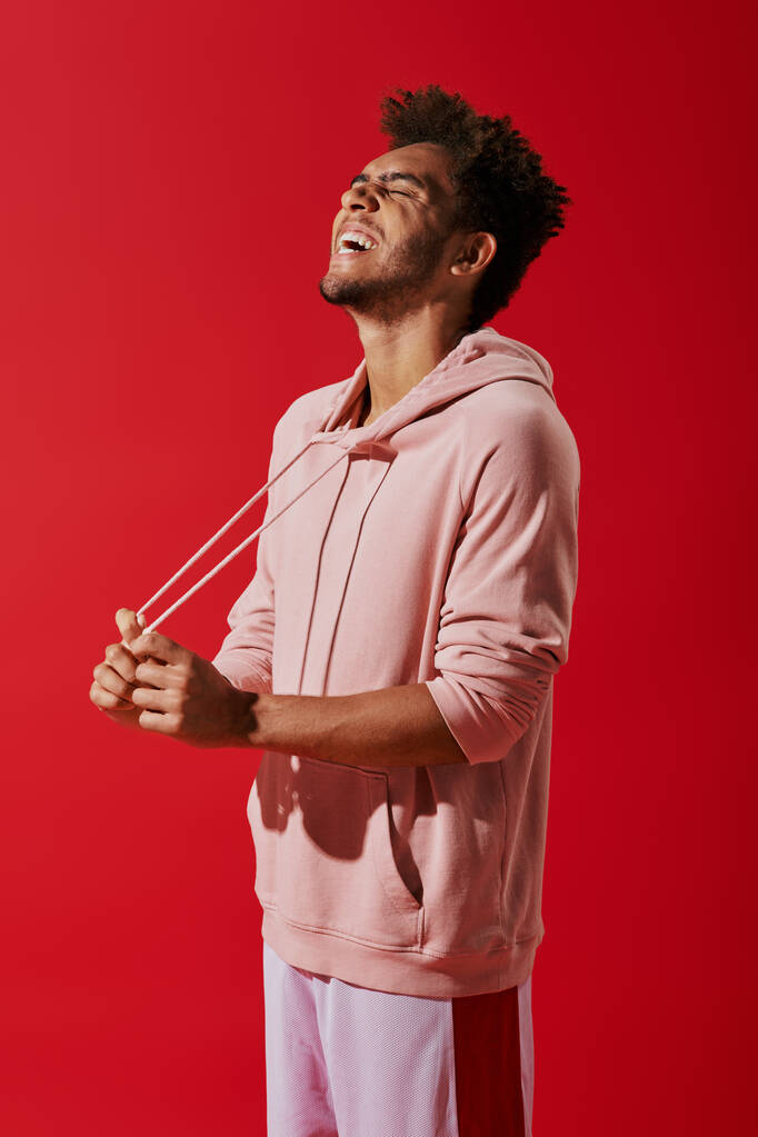 щасливий афроамериканський чоловік тягне струни рожевого светру і сміється на червоному тлі - Фото, зображення