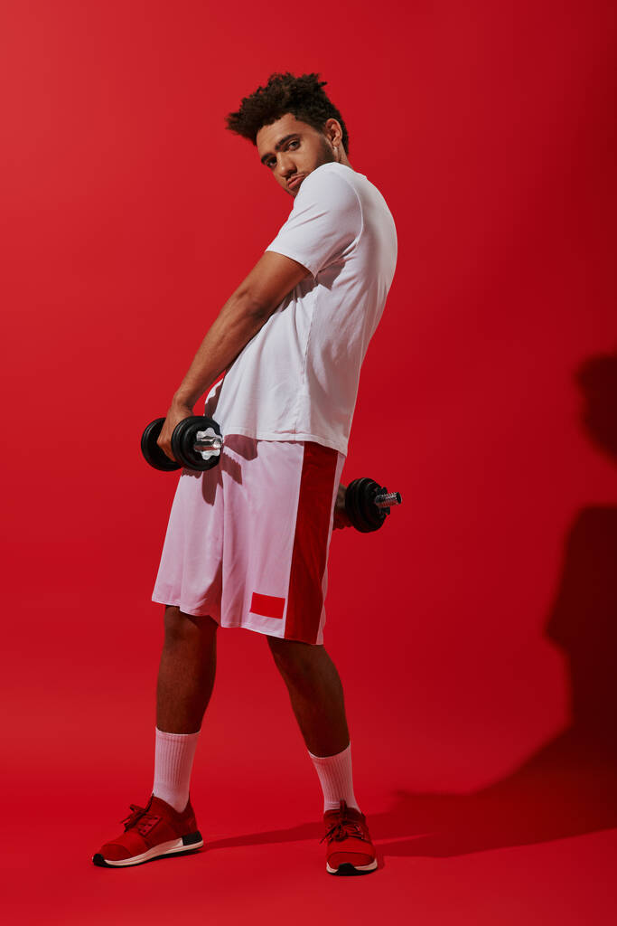 赤い背景で重いダンベルで運動するアフリカ系アメリカ人のスポーツマン,パワー - 写真・画像