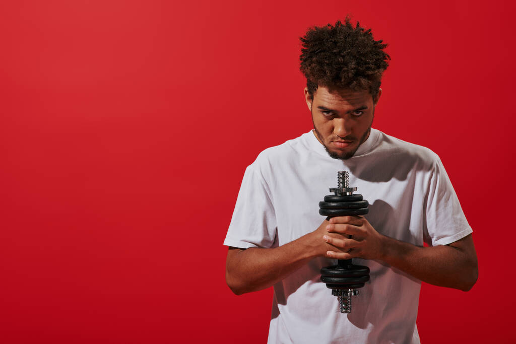 uomo africano americano in abbigliamento sportivo che lavora con manubri e guardando la fotocamera su sfondo rosso - Foto, immagini