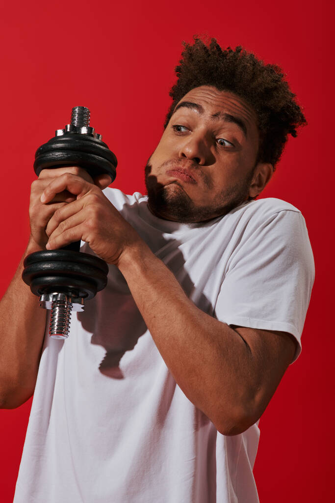 młody Afroamerykanin człowiek pracuje z ciężkimi hantlami i puffing policzki na czerwonym tle - Zdjęcie, obraz