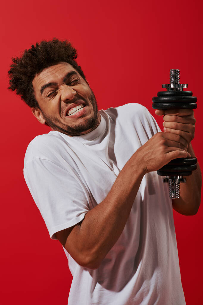 young and curly african american sportsman working out with heavy dumbbell on red background - Photo, Image