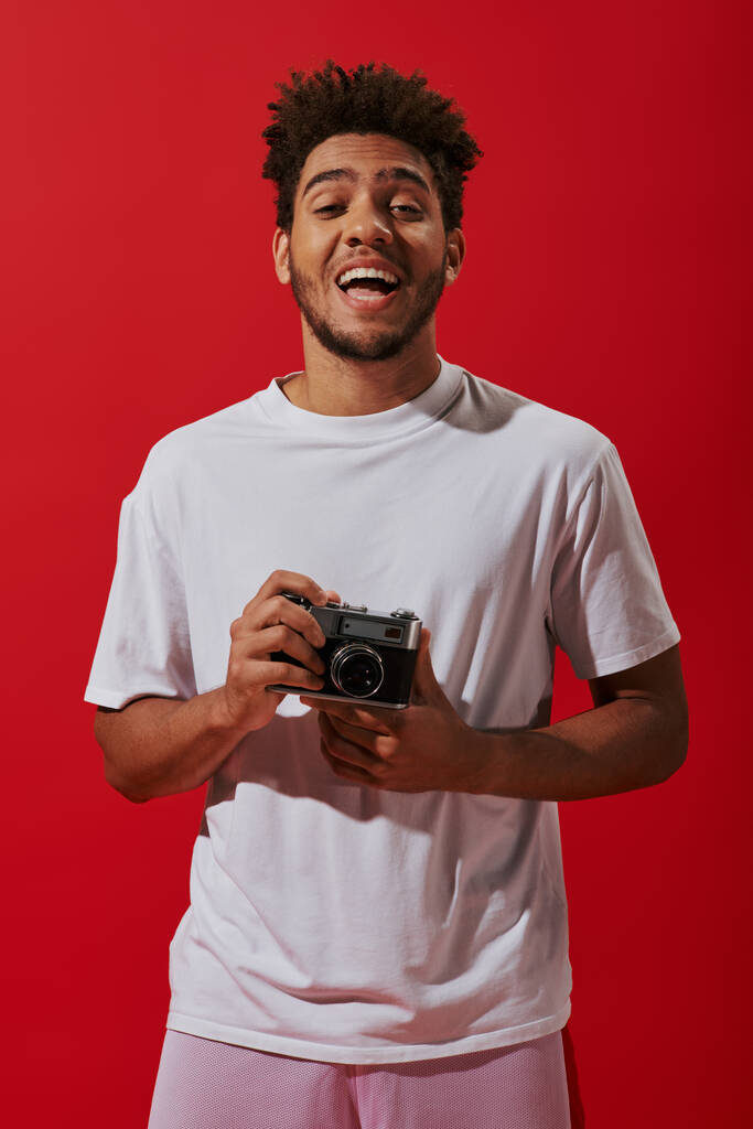 szczęśliwy afrykański fotograf trzyma zabytkowy aparat fotograficzny i uśmiecha się na czerwonym tle, hobby - Zdjęcie, obraz