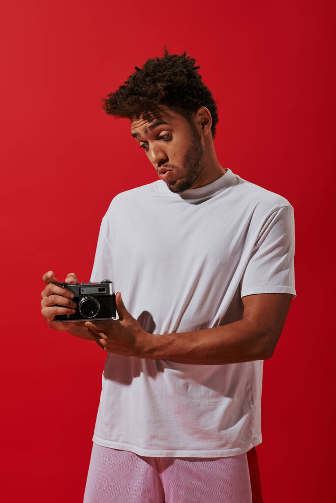 赤い背景の彼のヴィンテージカメラを見ている若いアフリカ系アメリカ人男性,趣味としての写真 - 写真・画像