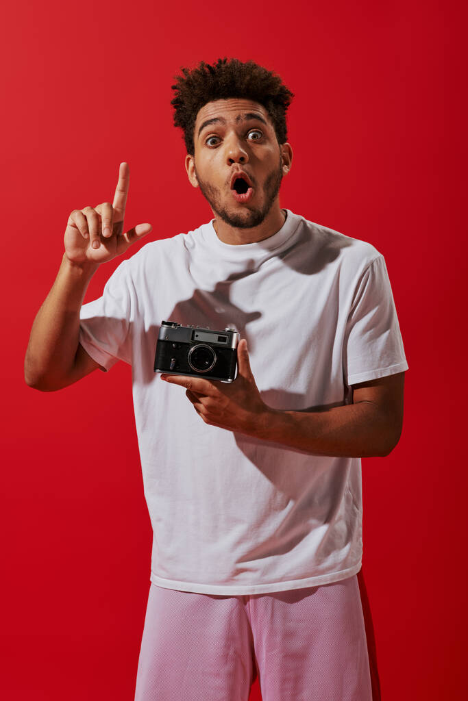 jeune homme afro-américain ayant idée tout en tenant son appareil photo vintage sur fond rouge - Photo, image