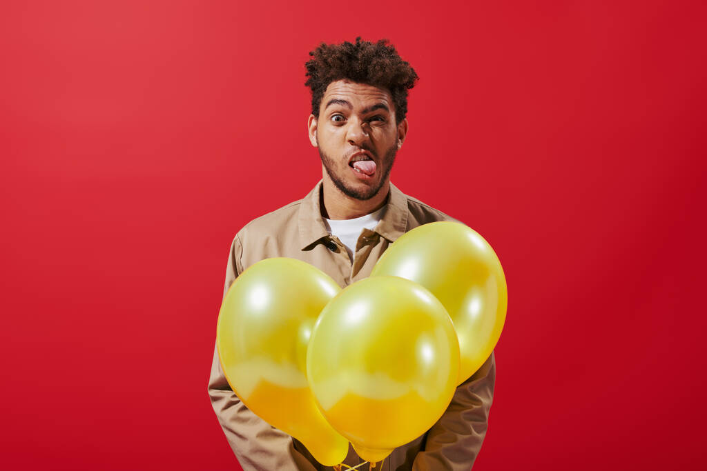 funny african american man in beige jacket holding balloons and sticking tongue on red background - Photo, Image