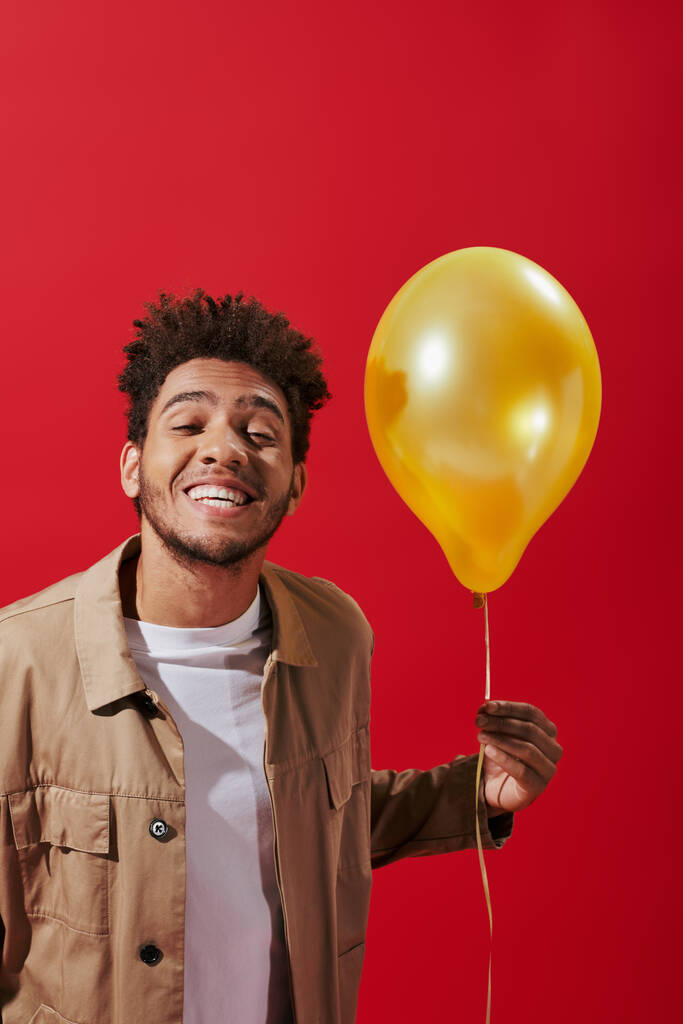 hombre afroamericano positivo en chaqueta beige sosteniendo globo y sonriendo sobre fondo rojo - Foto, imagen