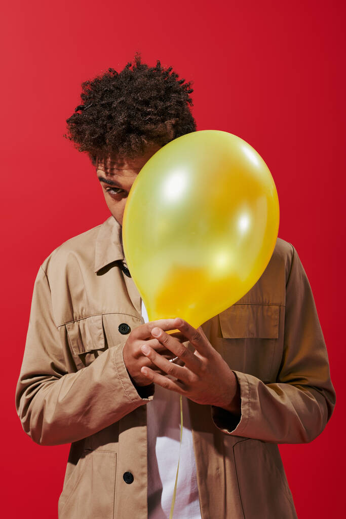 joven afroamericano hombre en chaqueta beige escondido detrás de globo sobre fondo rojo, oscureciendo la cara - Foto, Imagen