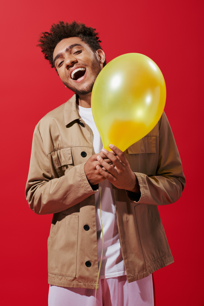 excited african american man in beige jacket holding balloon and smiling on red background - Photo, Image