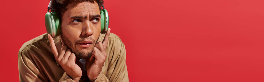 striscione di confuso uomo afroamericano in cuffie wireless ascoltare musica su sfondo rosso - Foto, immagini