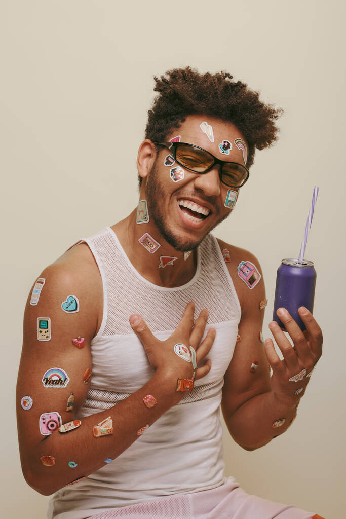 heureux homme afro-américain avec des autocollants sur le visage et le corps tenant canette de soda sur fond gris - Photo, image