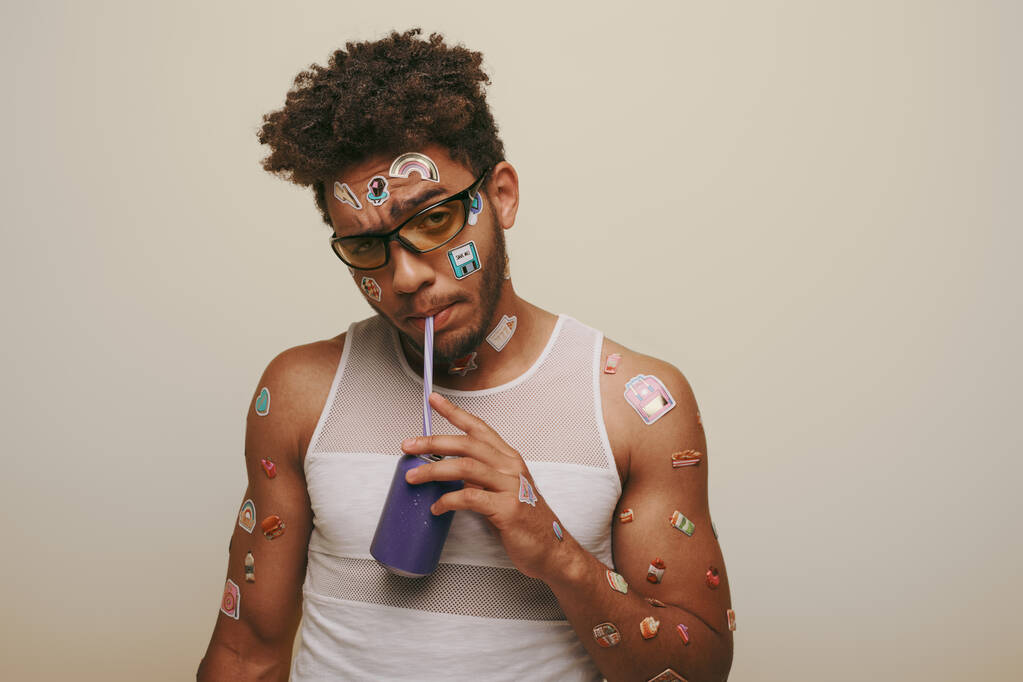 jovem afro-americano cara com adesivos no rosto e corpo bebendo refrigerante no fundo cinza - Foto, Imagem