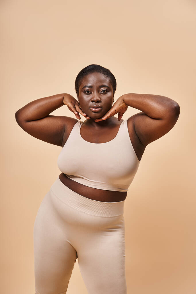 Plus size African American woman in beige underwear posing with hands near face on beige background - Photo, Image