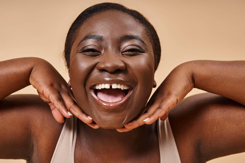 allegro plus size donna africana americana sorridente con le mani vicino al viso su sfondo beige - Foto, immagini