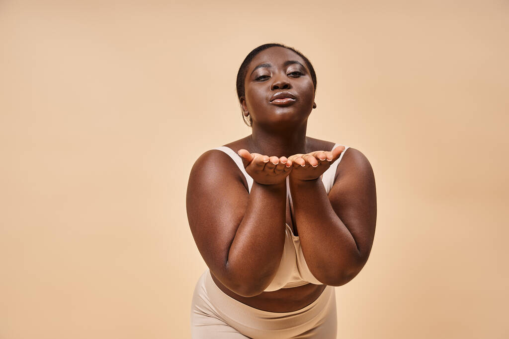 young plus size african american woman in beige underwear blowing air kiss against matching backdrop - Photo, Image