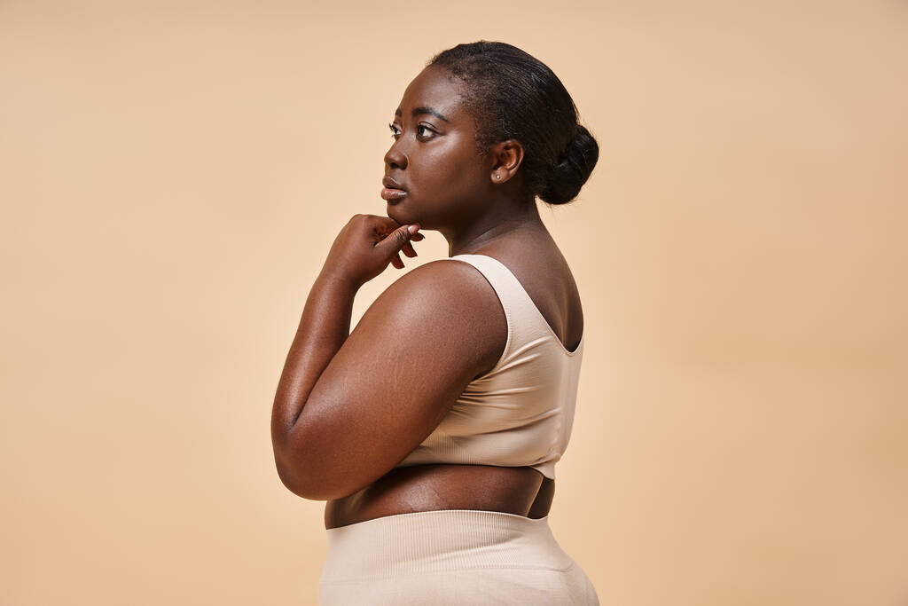 pensive plus size Afrikaans amerikaanse vrouw in beige ondergoed poseren tegen bijpassende achtergrond - Foto, afbeelding