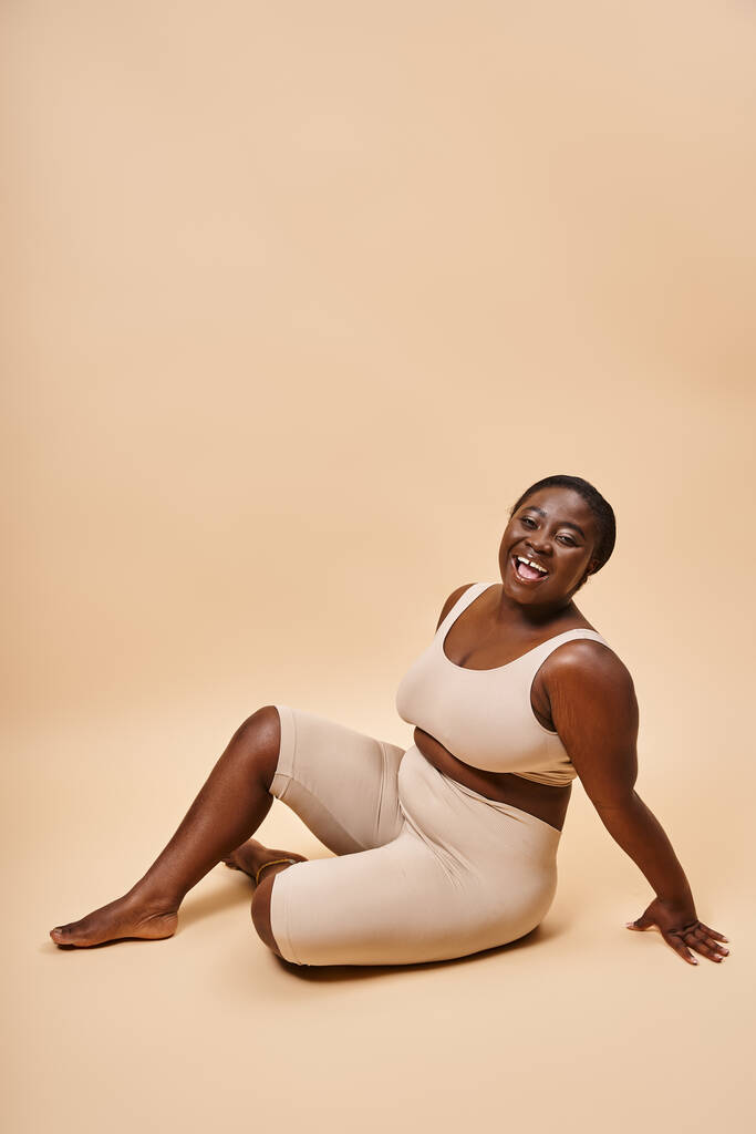 щасливий плюс розмір афроамериканської молодої жінки в бежевій білизні позує на відповідний фон - Фото, зображення