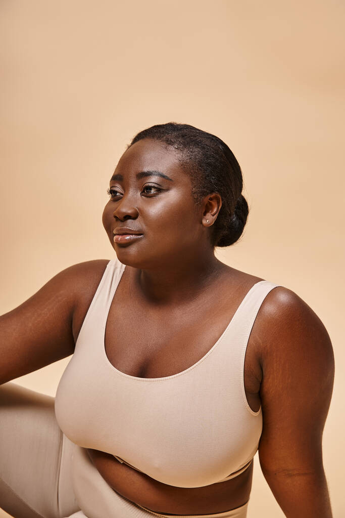 dreamy plus size african american young woman in beige underwear posing against matching backdrop - Photo, Image