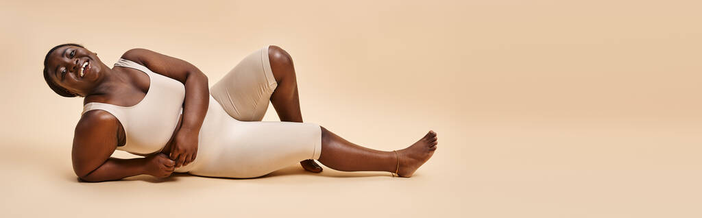 bannière de heureuse plus la taille afro-américaine jeune femme inclinant sur fond beige - Photo, image