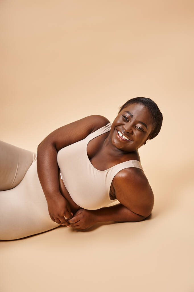 szczęśliwy plus rozmiar afrykański młody kobieta w beżowy bielizna leżące na dopasowanie tła - Zdjęcie, obraz
