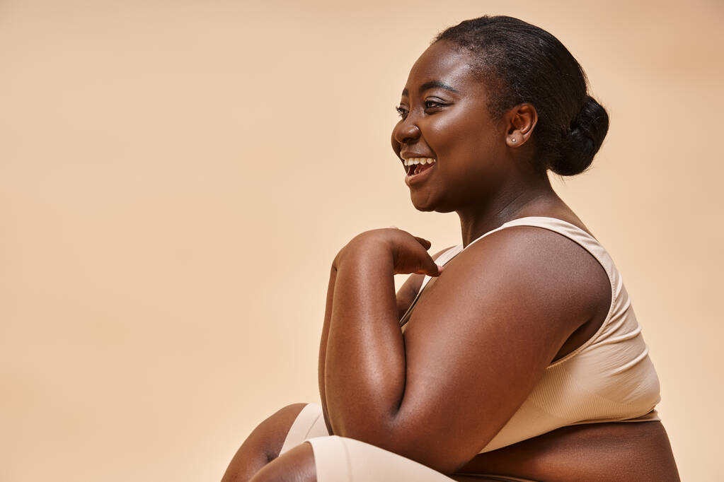 plus size modello afroamericano ridere e seduto sullo sfondo beige, corpo positivo - Foto, immagini