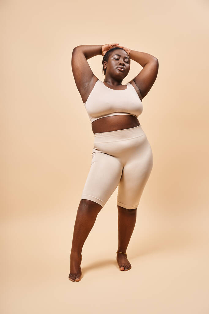 plus size Afrikaans Amerikaans model in ondergoed staand met opgeheven handen tegen beige achtergrond - Foto, afbeelding