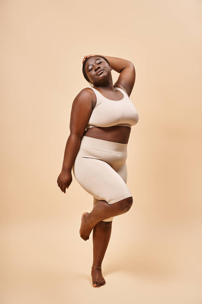 más tamaño mujer afroamericana en ropa interior de pie con la mano levantada contra el telón de fondo beige - Foto, imagen