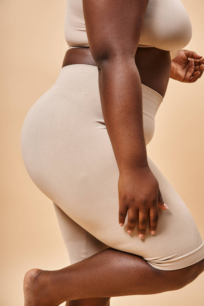 обрезанный вид плюс размер африканской американки в нижнем белье позирует на бежевом фоне, пышное тело - Фото, изображение