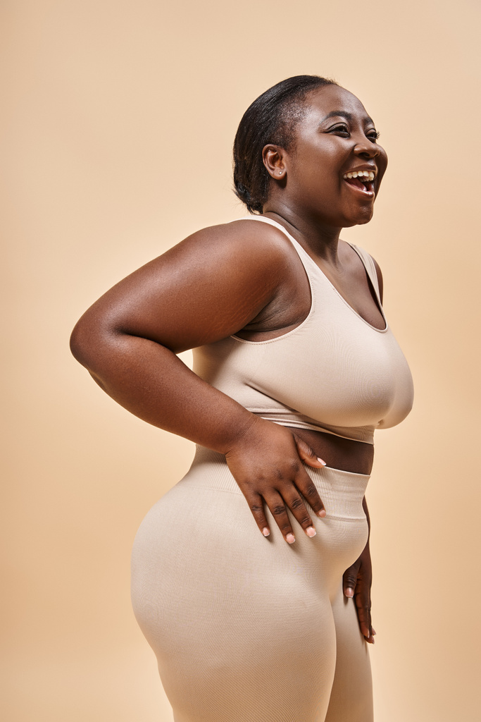 Vreugdevolle Afrikaanse Amerikaanse plus size vrouw in beige ondergoed omarmen zelf-liefde en vertrouwen - Foto, afbeelding