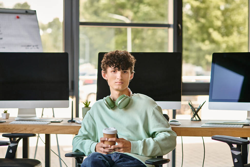 jeune homme aux cheveux bouclés assis dans une chaise de bureau et tenant un café, membre de l'équipe de post-production - Photo, image