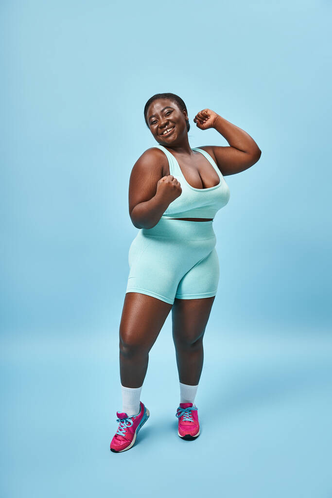 aufgeregte Plus Size Frau in aktivem Tragen lässt Muskeln spielen und blickt in die Kamera auf blauem Hintergrund - Foto, Bild