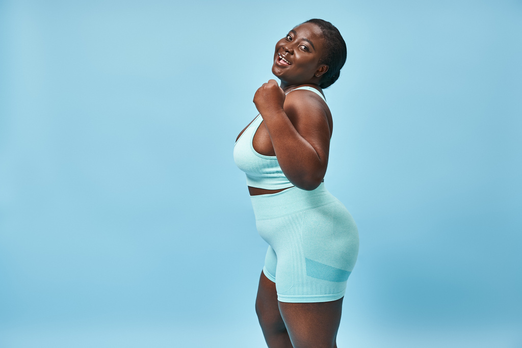 happy plus size woman in active wear flexing her muscles and looking at camera on blue background - Photo, Image