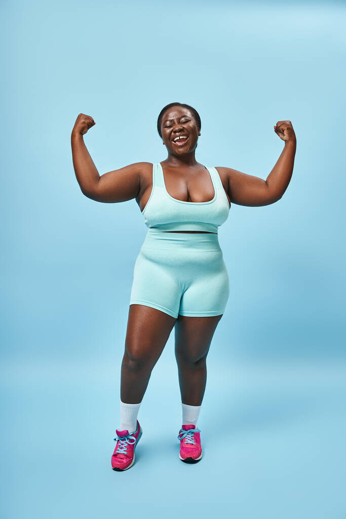 excited plus size woman in active wear flexing her muscles and smiling on blue background - Photo, Image