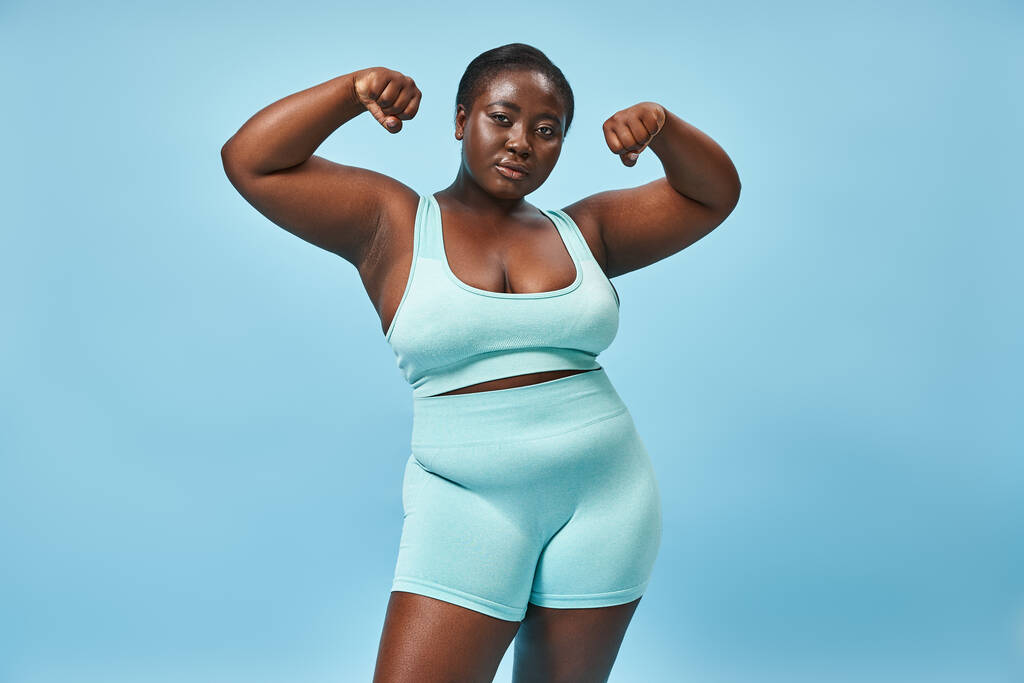vážná plus velikost žena v aktivním oblečení ohýbání svaly a při pohledu na kameru na modrém pozadí - Fotografie, Obrázek