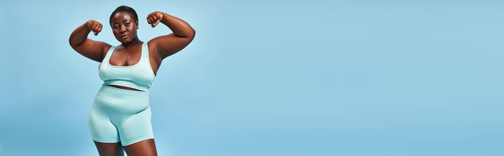 banner, plus size vrouw in actieve slijtage flexen haar spieren en kijken naar camera op blauwe achtergrond - Foto, afbeelding
