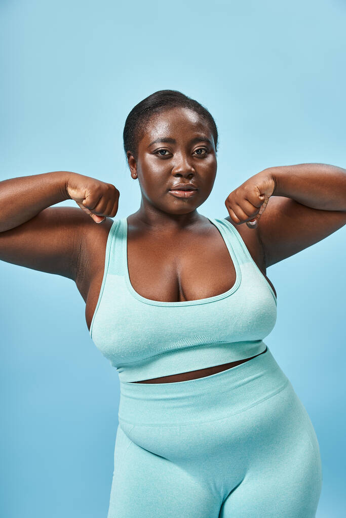 sport, plus size woman in active wear flexing her muscles and looking at camera on blue background - Photo, Image