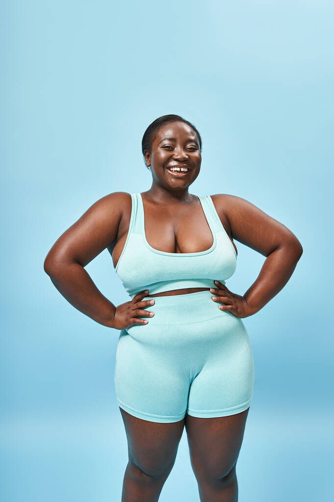 happy african american woman in blue sportswear with hands on hips on matching backdrop - Photo, Image