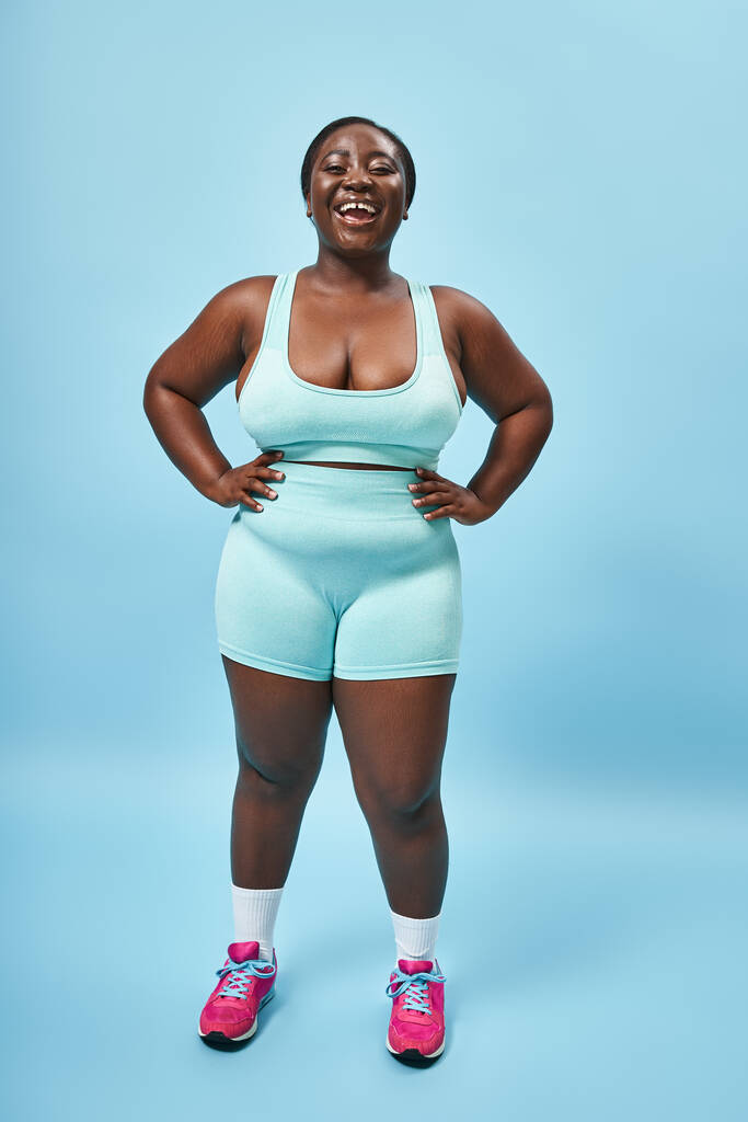 jolly plus size donna africana americana in blu abbigliamento sportivo con le mani sui fianchi sullo sfondo corrispondente - Foto, immagini