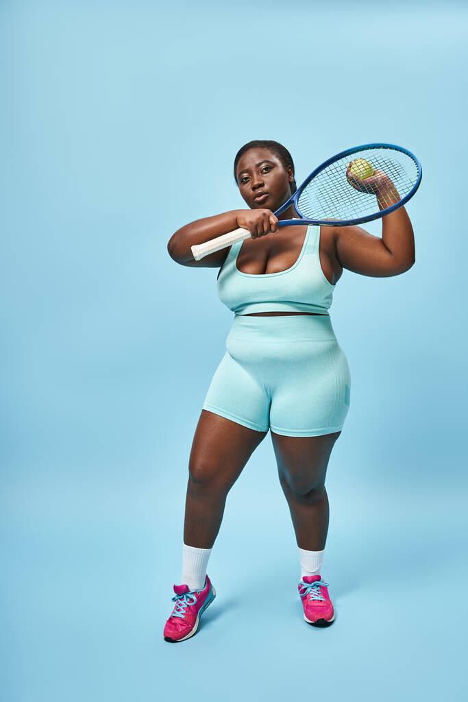 αυτοπεποίθηση συν μέγεθος αφροαμερικανή γυναίκα έτοιμη να παίξει τένις σε μπλε φόντο, το σώμα θετικό - Φωτογραφία, εικόνα
