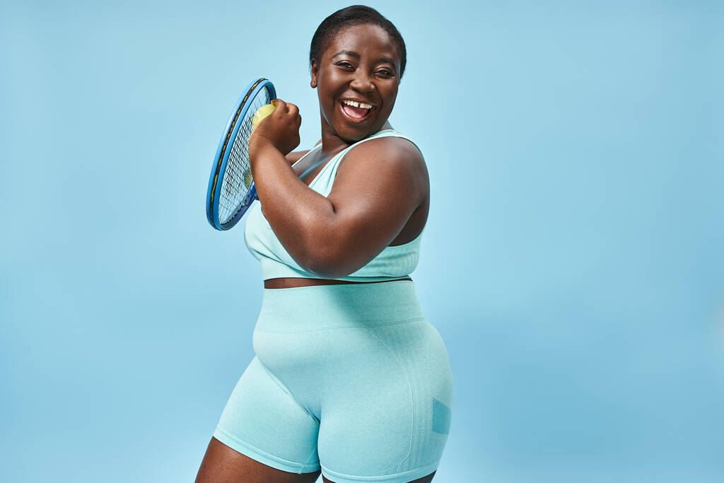 vidám plusz méret afro-amerikai nő kész teniszezni kék háttér, test pozitív - Fotó, kép