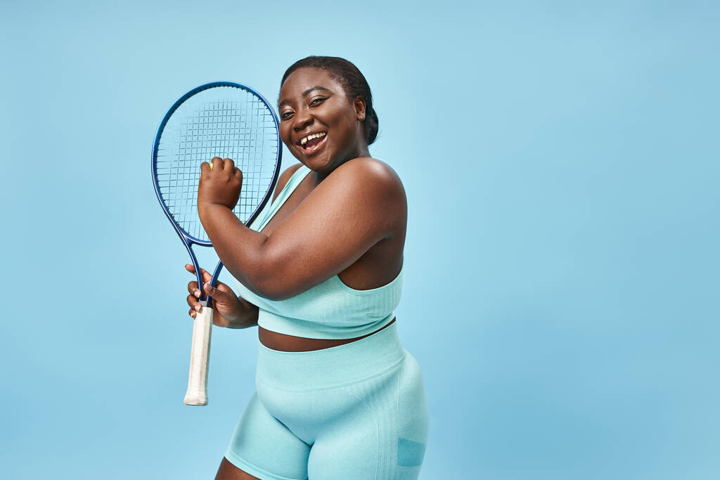 χαμογελαστός συν μέγεθος αφροαμερικανή γυναίκα έτοιμη να παίξει τένις σε μπλε φόντο, το σώμα θετικό - Φωτογραφία, εικόνα
