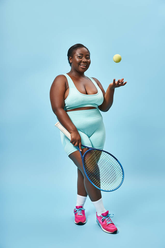veselý plus velikost africké americká žena drží tenisovou raketu a házení míč na modrém pozadí - Fotografie, Obrázek