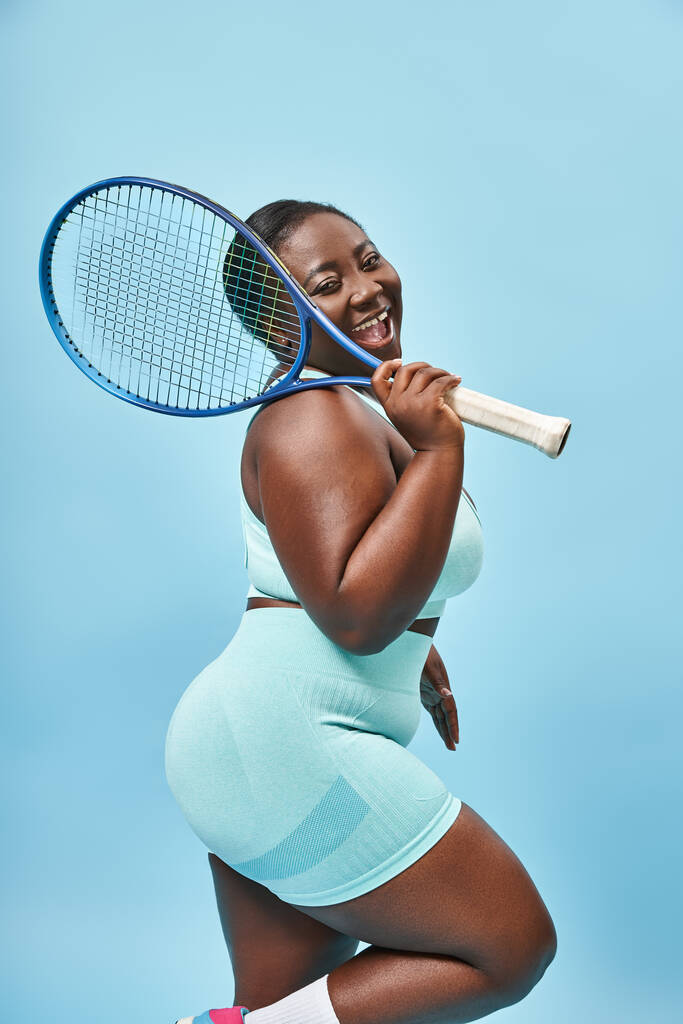 jolly plus size dark skinned woman holding tennis racket on blue backdrop, body positive and sport - Photo, Image