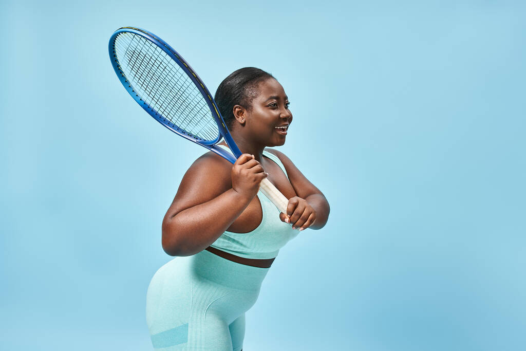 radiante más tamaño de piel oscura mujer sosteniendo raqueta de tenis sobre fondo azul, cuerpo positivo y deporte - Foto, imagen