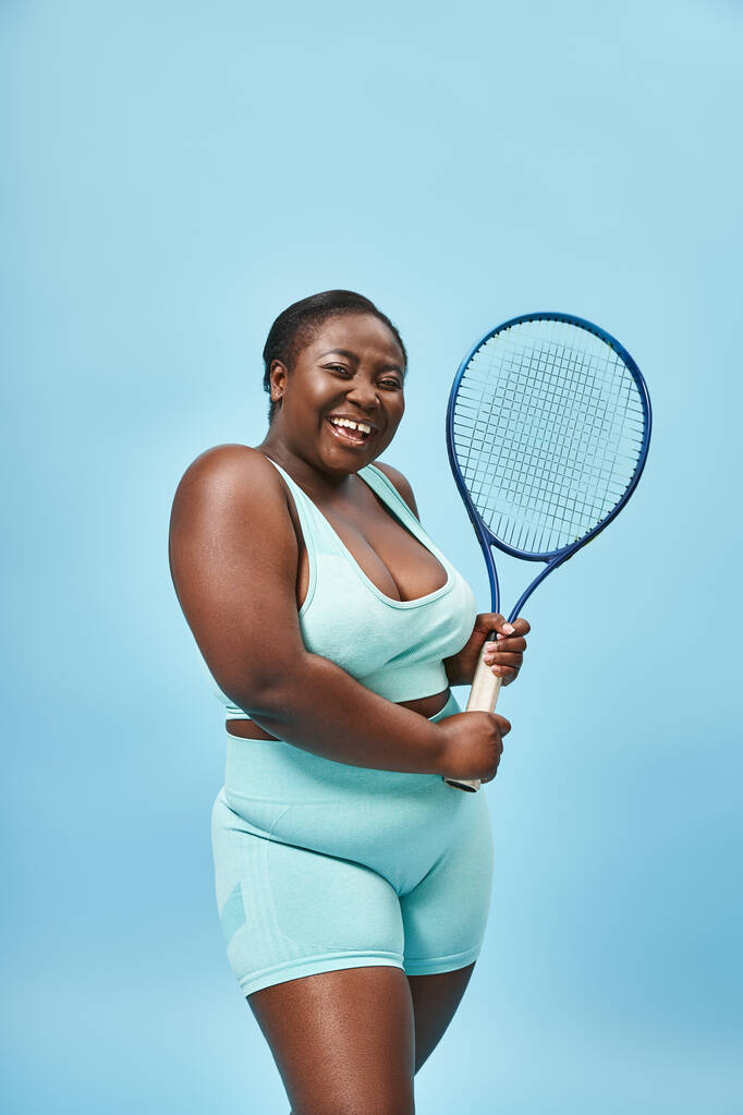 ευτυχισμένη συν μέγεθος μαύρη γυναίκα κρατώντας ρακέτα τένις σε μπλε φόντο, το σώμα θετικό και τον αθλητισμό - Φωτογραφία, εικόνα