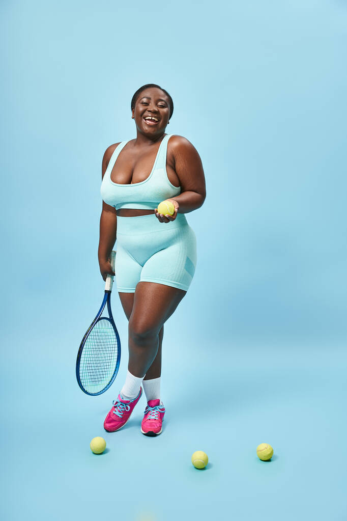 alegre más tamaño de piel oscura deportista sosteniendo raqueta de tenis y pelota sobre fondo azul, jugador - Foto, Imagen