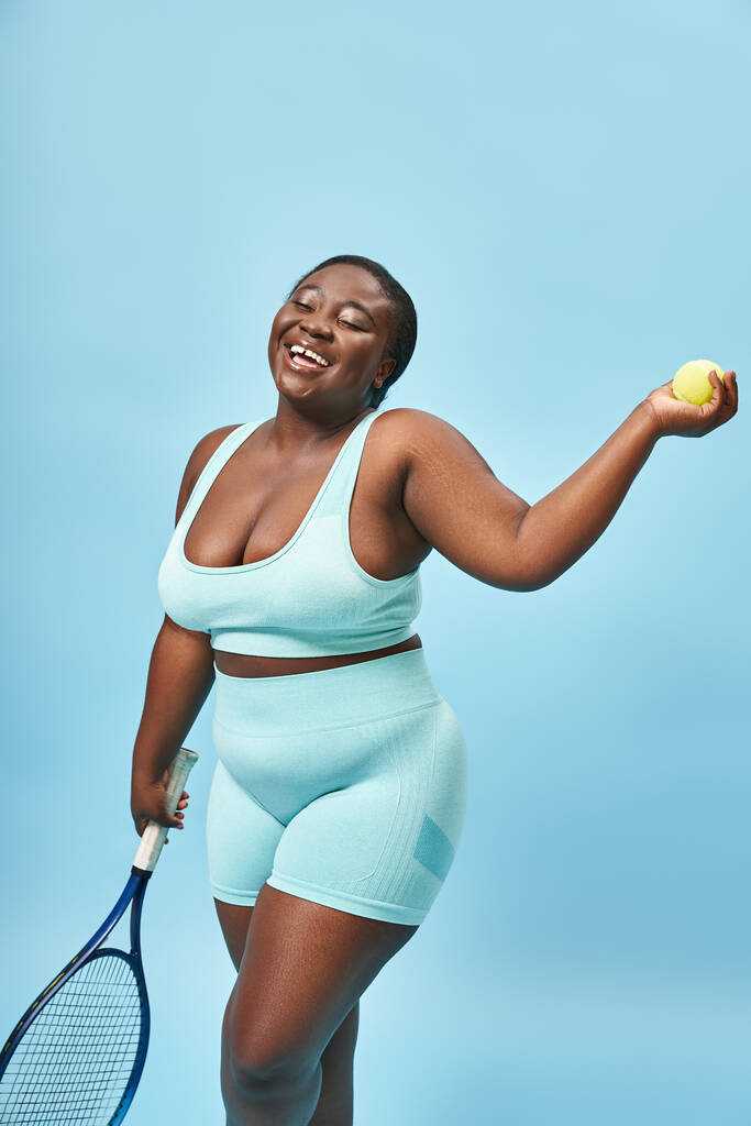 glimlachen plus size donkere huid sporter met tennis racket en bal op blauwe achtergrond, speler - Foto, afbeelding