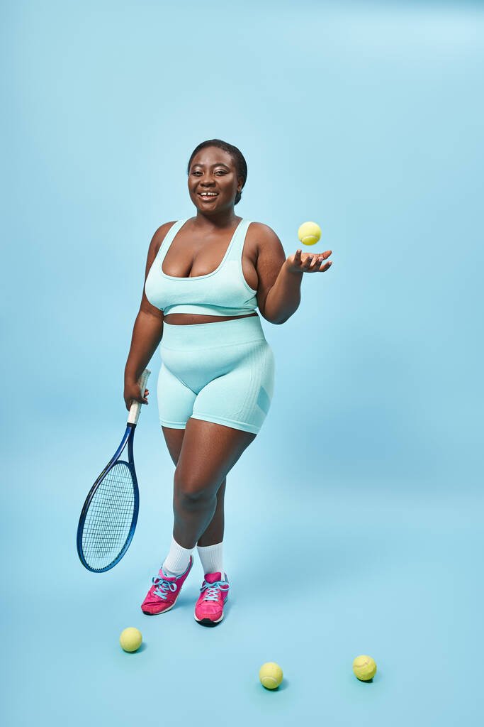 mladý plus velikost tmavé kůže sportovkyně drží tenisovou raketu a míč na modrém pozadí, hráč - Fotografie, Obrázek