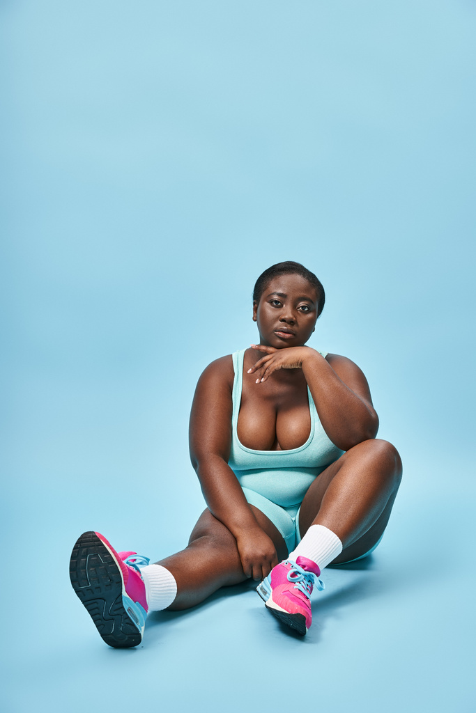 Relajado más tamaño afroamericana deportista sentado en ropa deportiva azul en el telón de fondo a juego - Foto, Imagen