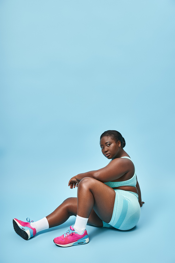 Relajada mujer afroamericana de tamaño grande sentada en ropa deportiva azul en el telón de fondo a juego, deporte - Foto, imagen